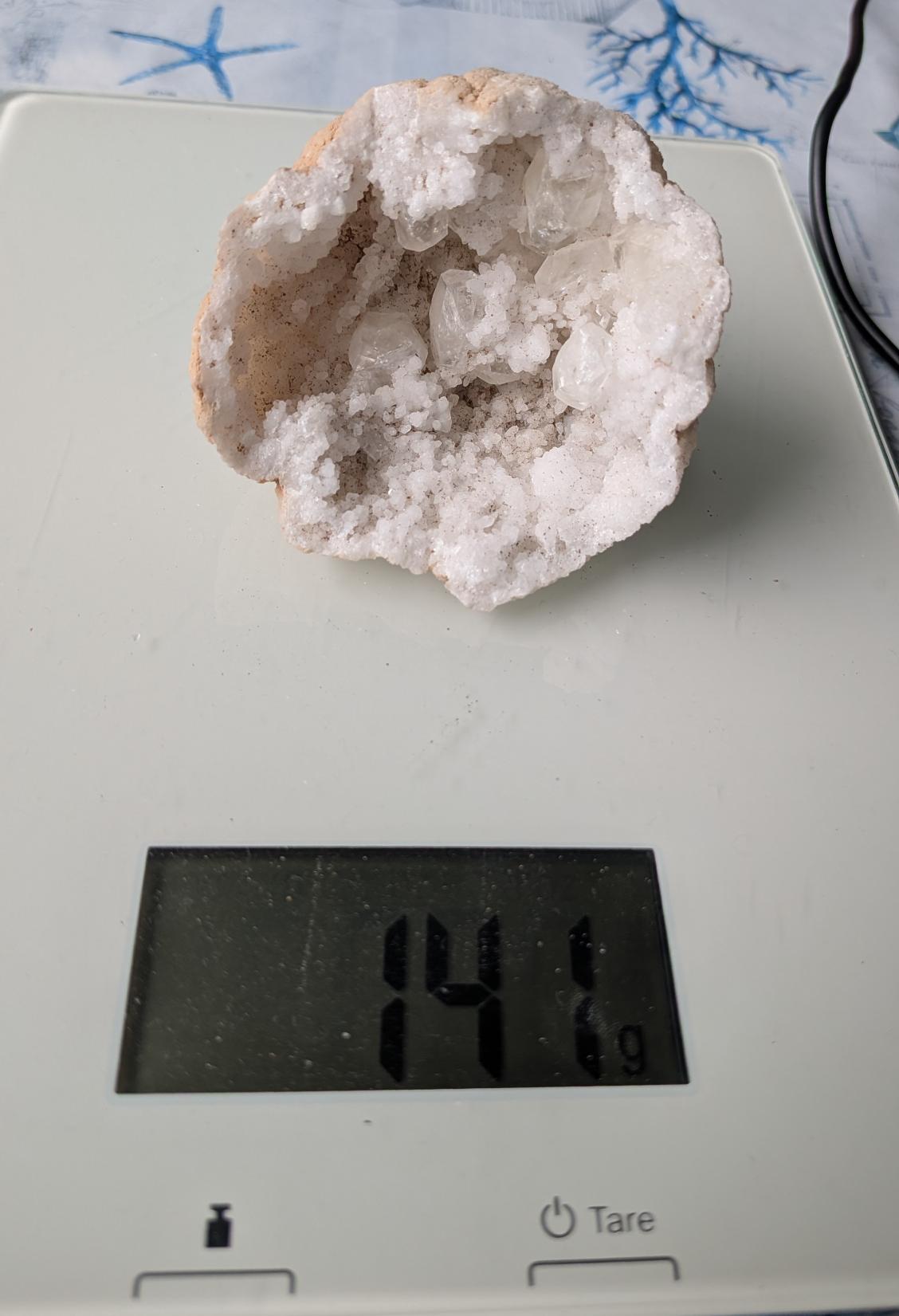 Een natuurlijke Bergkristal geode van 141 gram met fonkelende, heldere kristalpunten aan de binnenzijde, gefotografeerd op een digitale weegschaal.