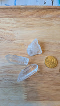 Drie heldere ruwe bergkristallen, waaronder twee kristalpunten, naast een 50 eurocent munt op een houten ondergrond voor een custom wire wrapped hanger.