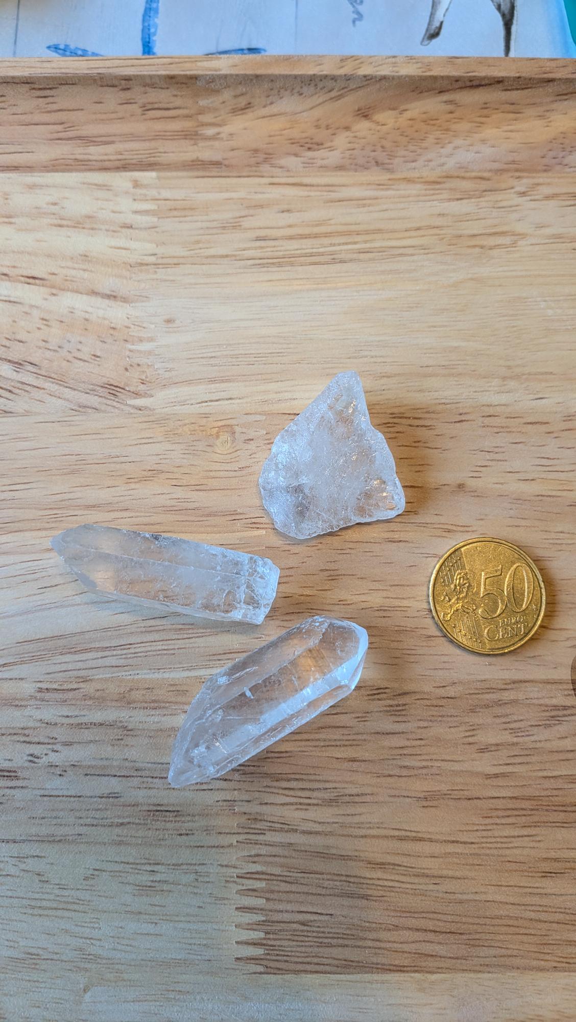 Drie heldere ruwe bergkristallen, waaronder twee kristalpunten, naast een 50 eurocent munt op een houten ondergrond voor een custom wire wrapped hanger.