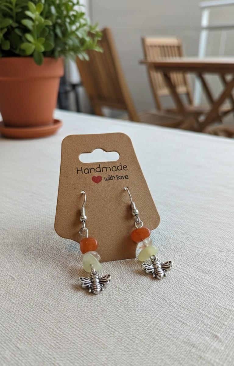 Handgemaakte zilverkleurige bijen-oorbellen met oranje en groene edelsteen kralen op een kraft kaartje.