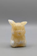 Handgeslepen edelsteen figuurtje van een zittende gele Pokémon (Pikachu), geslepen uit Gele Calciet met een zachte glans, tegen een grijze achtergrond.