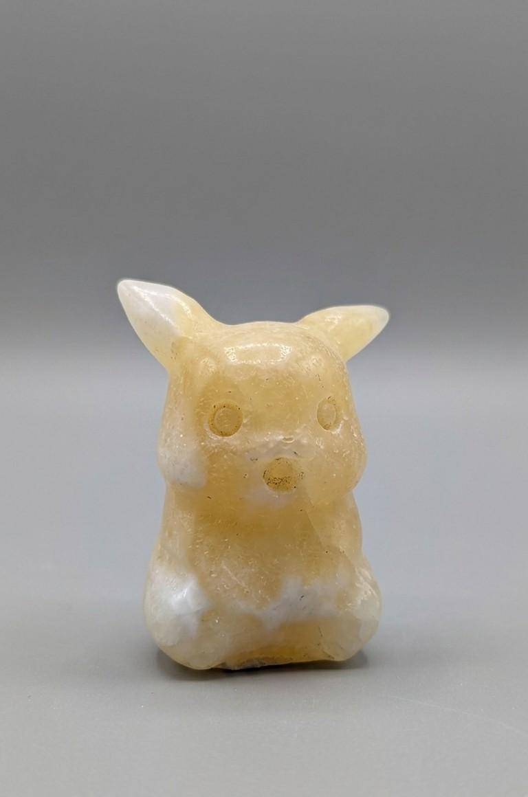 Handgeslepen edelsteen figuurtje van een zittende gele Pokémon (Pikachu), geslepen uit Gele Calciet met een zachte glans, tegen een grijze achtergrond.