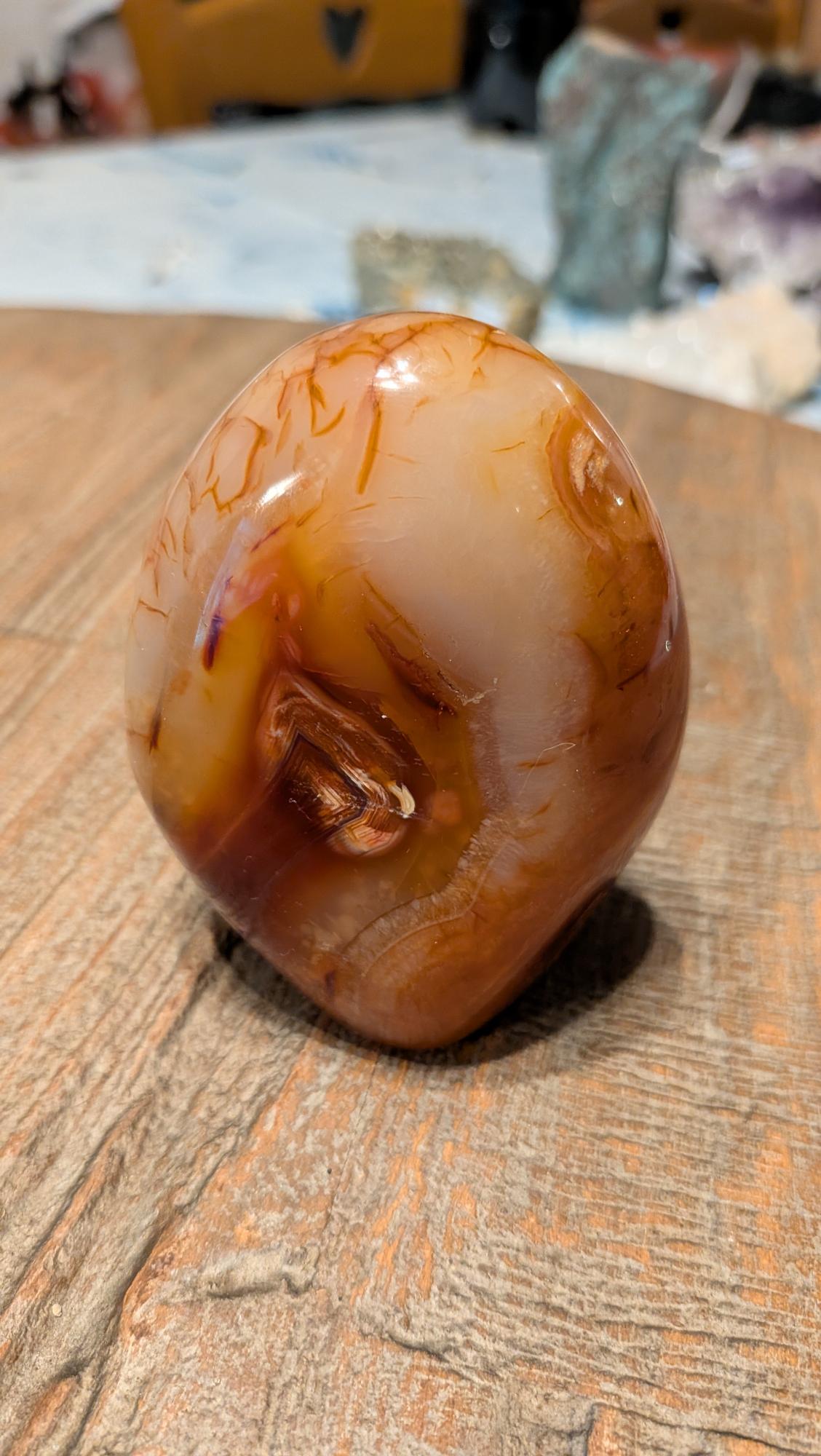 Gepolijste oranje Carneool freeform van 264 gram met natuurlijke bandenstructuur op een houten ondergrond.