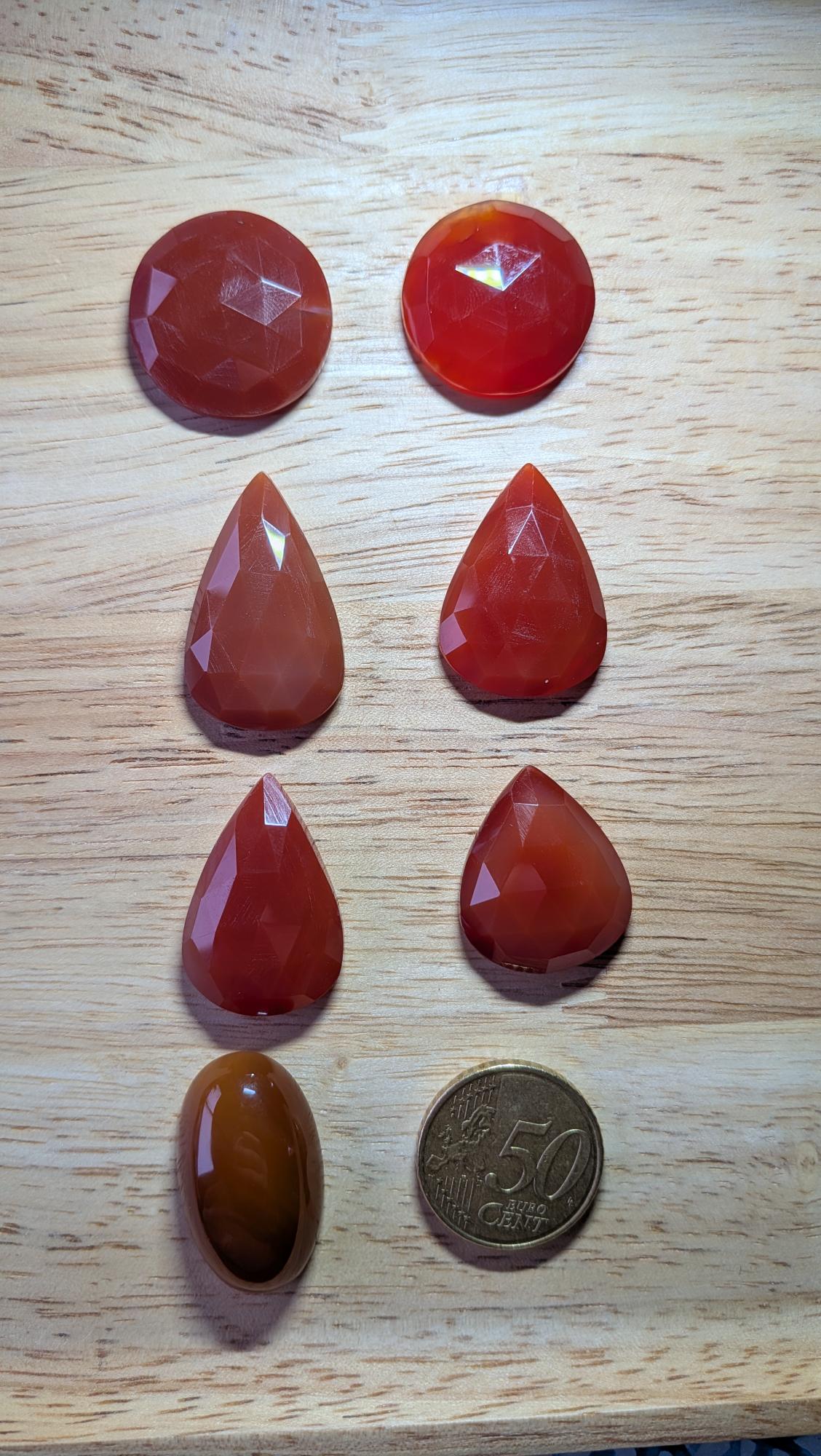 Zeven oranje Carneool edelstenen in diverse vormen, waaronder facetgeslepen druppels en een gladde ovale cabochon, naast een 50 cent muntstuk.