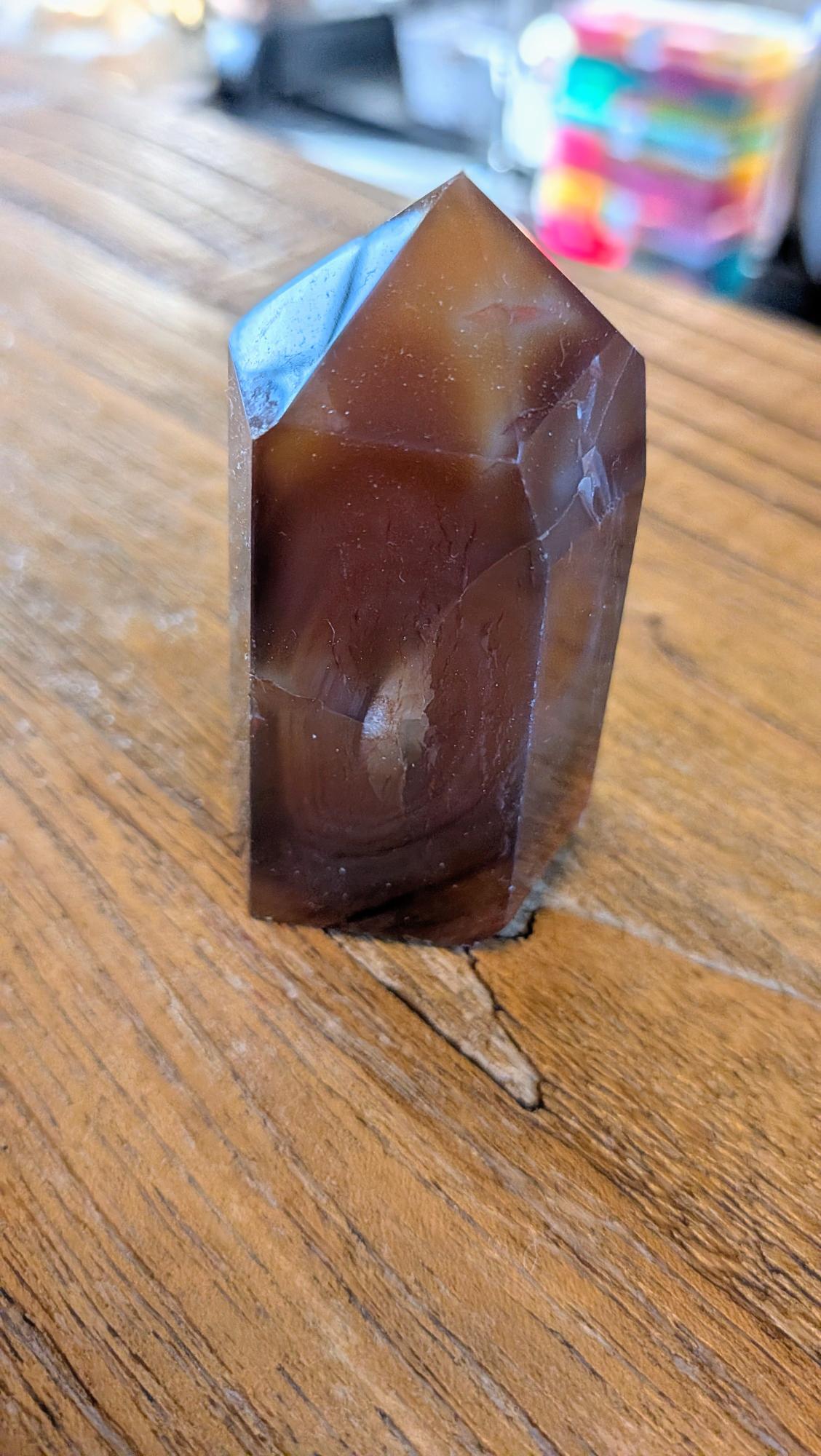 "Donkeroranje Carneool kristal tower met een opvallende inkeping/beschadiging staand op een houten tafel."