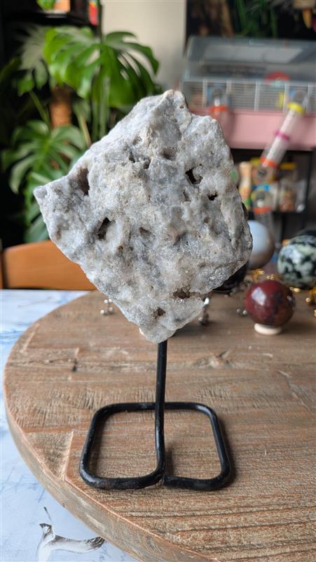 Voorzijde van een witte chalcedoon edelsteen op zwarte standaard, tonend een natuurlijke holte met fijne, fonkelende kristallen.