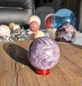 Paarse Chevron Amethist bol van 6 cm diameter en 317 gram met wit V-patroon op een rode houten standaard