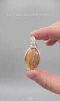 Handgemaakte wire wrapped hanger van Edeltwist met een Crazy Lace Agaat (lachsteen) in een zilveren wikkeling tegen een neutrale, grijze achtergrond.