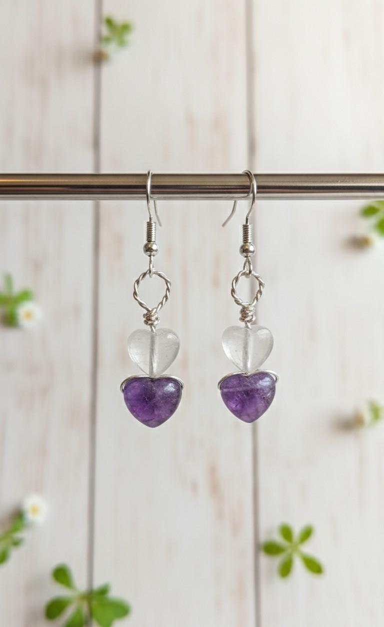 Handgemaakte zilverkleurige oorbellen met een helder bergkristal hartje boven een paars amethist hartje, hangend aan een metalen standaard.