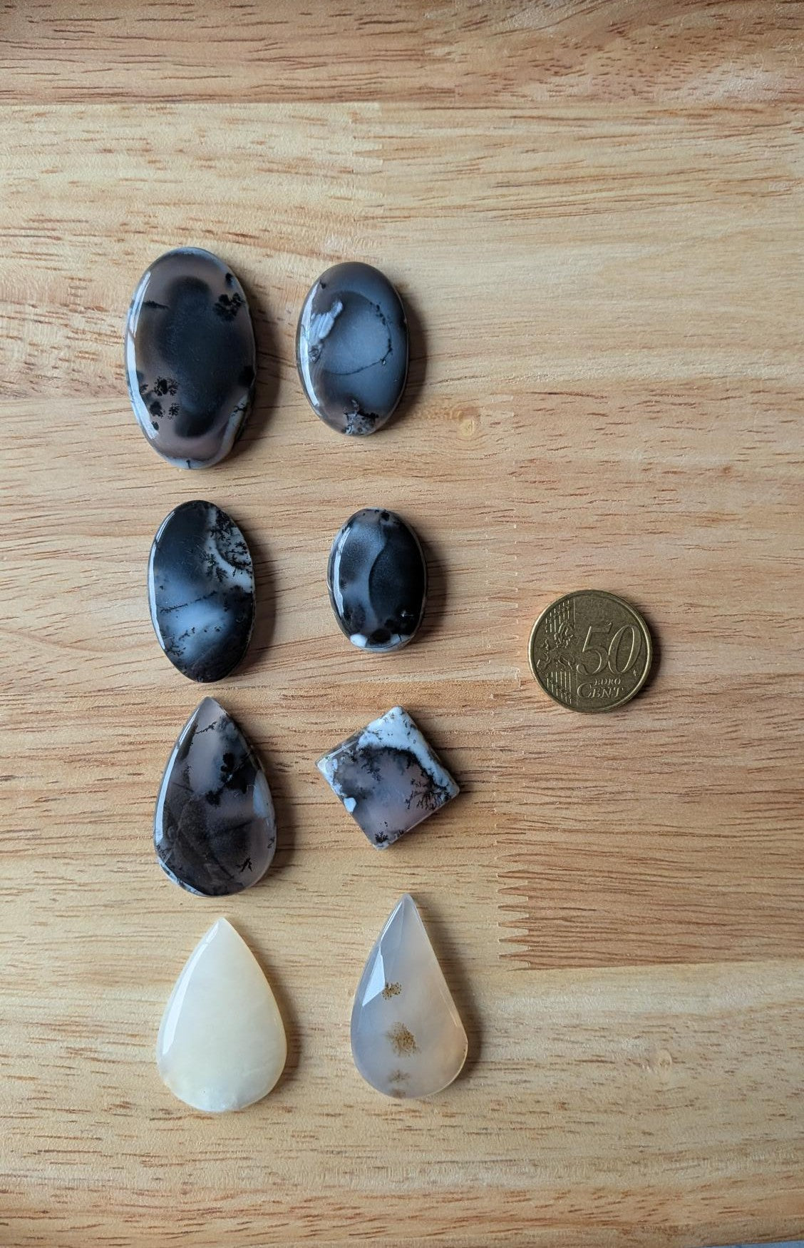 Acht verschillende Dendriet Agaat edelstenen met boomachtige insluitingen in vormen zoals ovaal, druppel en ruit op een houten achtergrond naast een 50 cent muntstuk.
