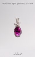 Een close-up detailopname van de unieke, handgemaakte zilveren wire-wrapped hanger met de opvallende magenta gekleurde drakenader agaatsteen. De hanger rust op een grijze vilten displaypad. De focus ligt op het ingewikkelde zilveren draadwerk, inclusief de minuscule spiralen en de complexe, handgemaakte knoopstructuur die de steen veilig vasthoudt en de bail vormt. De unieke interne patronen van de drakenader agaat, met zijn diepe magenta kleur en zwarte aders, zijn scherp en duidelijk zichtbaar. De hanger 