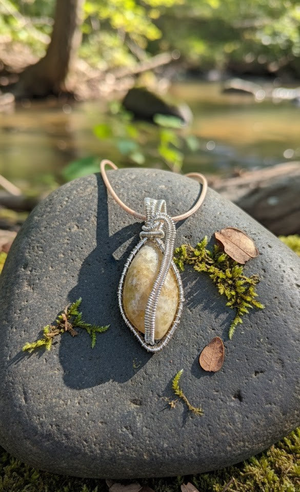 Handgemaakte verzilverde wire-wrapped hanger met een druppelvormige fossielkoraal edelsteen, liggend op een bemoste steen bij een kabbelend beekje in het bos.