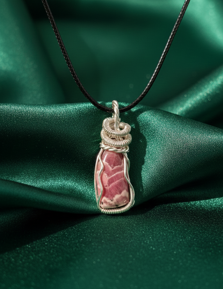 Handgemaakte zilverkleurige wire-wrapped hanger met een gepolijste roze Rhodochrosiet edelsteen, gepresenteerd op glanzend donkergroen satijn.