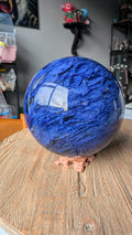 Een gigantische, diepblauwe smeltkwarts bol van 18 cm diameter en 6.85 kilo met een glanzend oppervlak en interne gelaagde structuren, stabiel gepresenteerd op een sierlijke, roodbruine houten houder op een rustieke tafel.