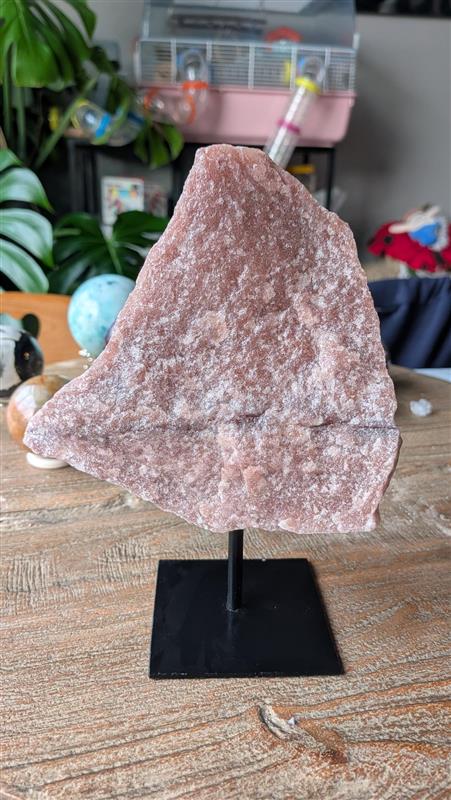 XL-formaat ruwe rozenkwarts edelsteen op zwarte standaard, gewicht 1042 gram en 19 cm hoog. De roze steen heeft een natuurlijke, robuuste vorm en staat op een houten ondergrond.