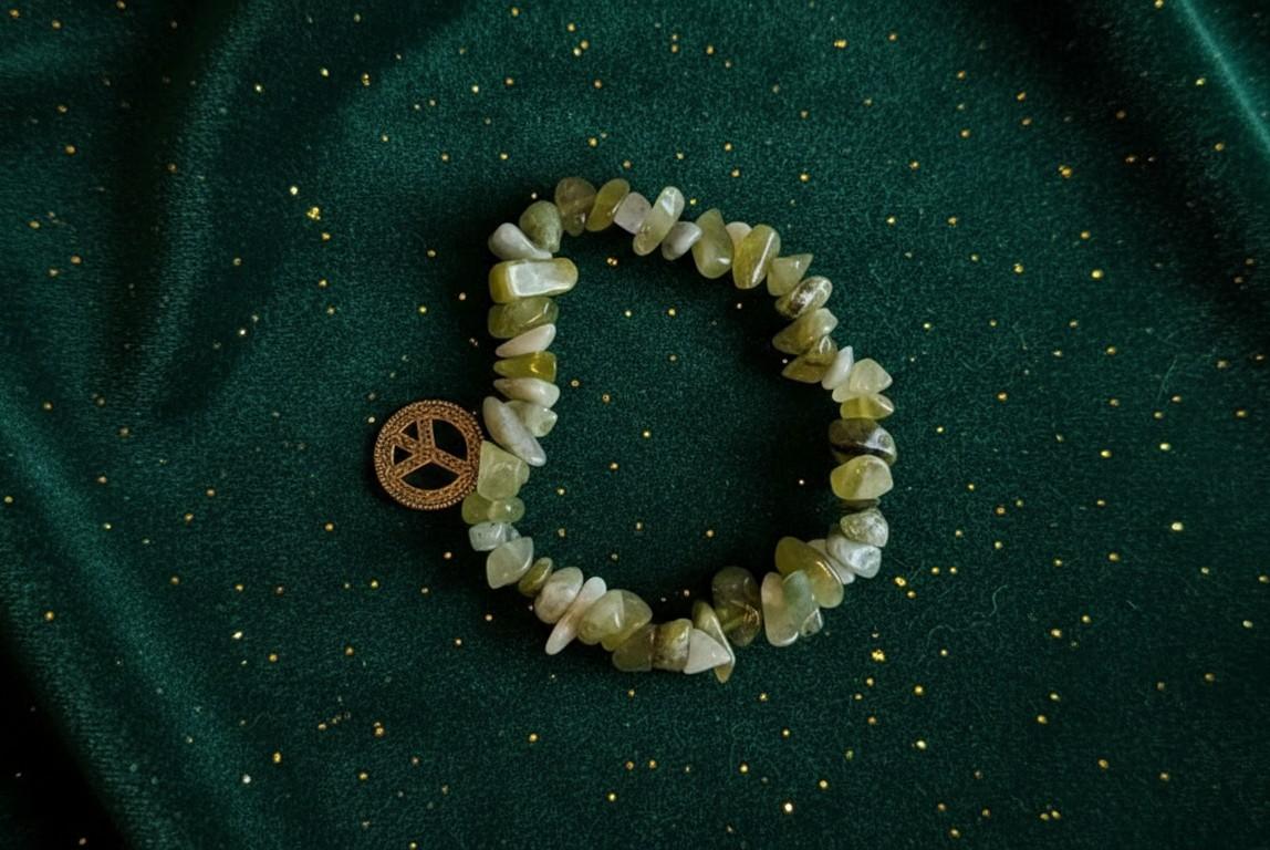 Handgemaakte armband van lichtgroene en gele edelsteen chips met een gouden peace-bedel op een donkergroene fluwelen achtergrond.