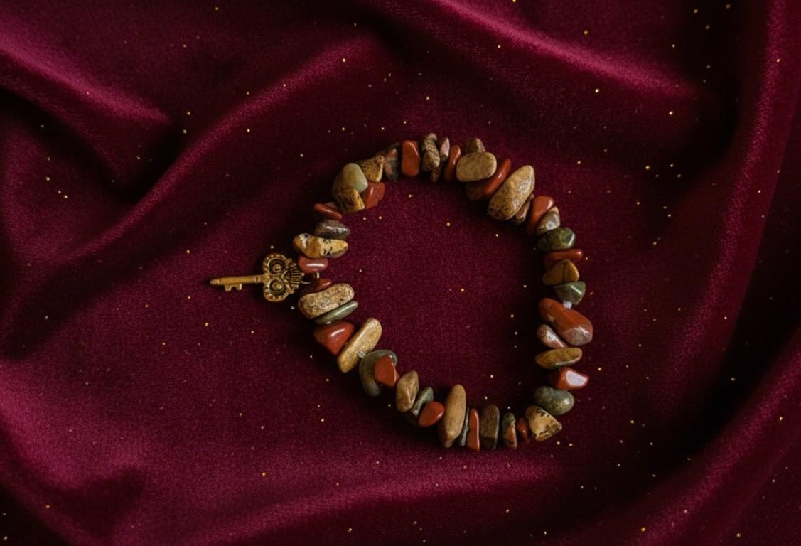 Handgemaakte natuursteen armband met rode jaspis en groene unakiet chips en een goudkleurige sleutel bedel op een bordeauxrode fluwelen achtergrond.