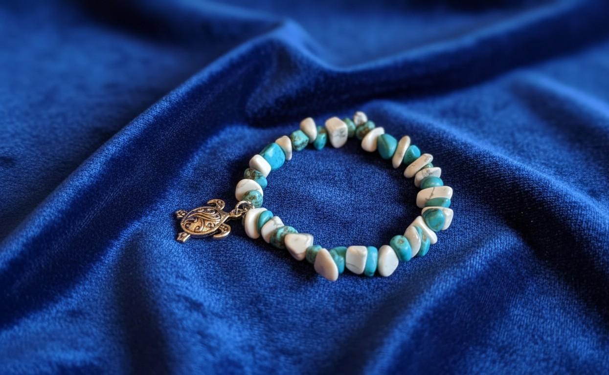 Handgemaakte natuursteen armband met witte Howliet en blauwe Turkoois chips en een zilveren schildpad bedel op een blauwe fluwelen achtergrond.