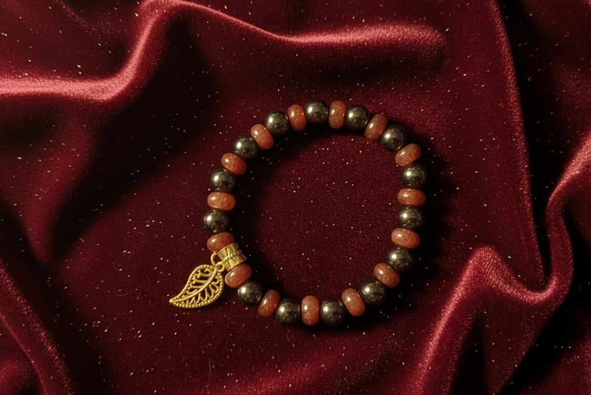 Handgemaakte armband van warme terra-kleurige zonnesteen en metallic grijze hematiet kralen met een gouden blad-bedel op een bordeauxrode achtergrond.