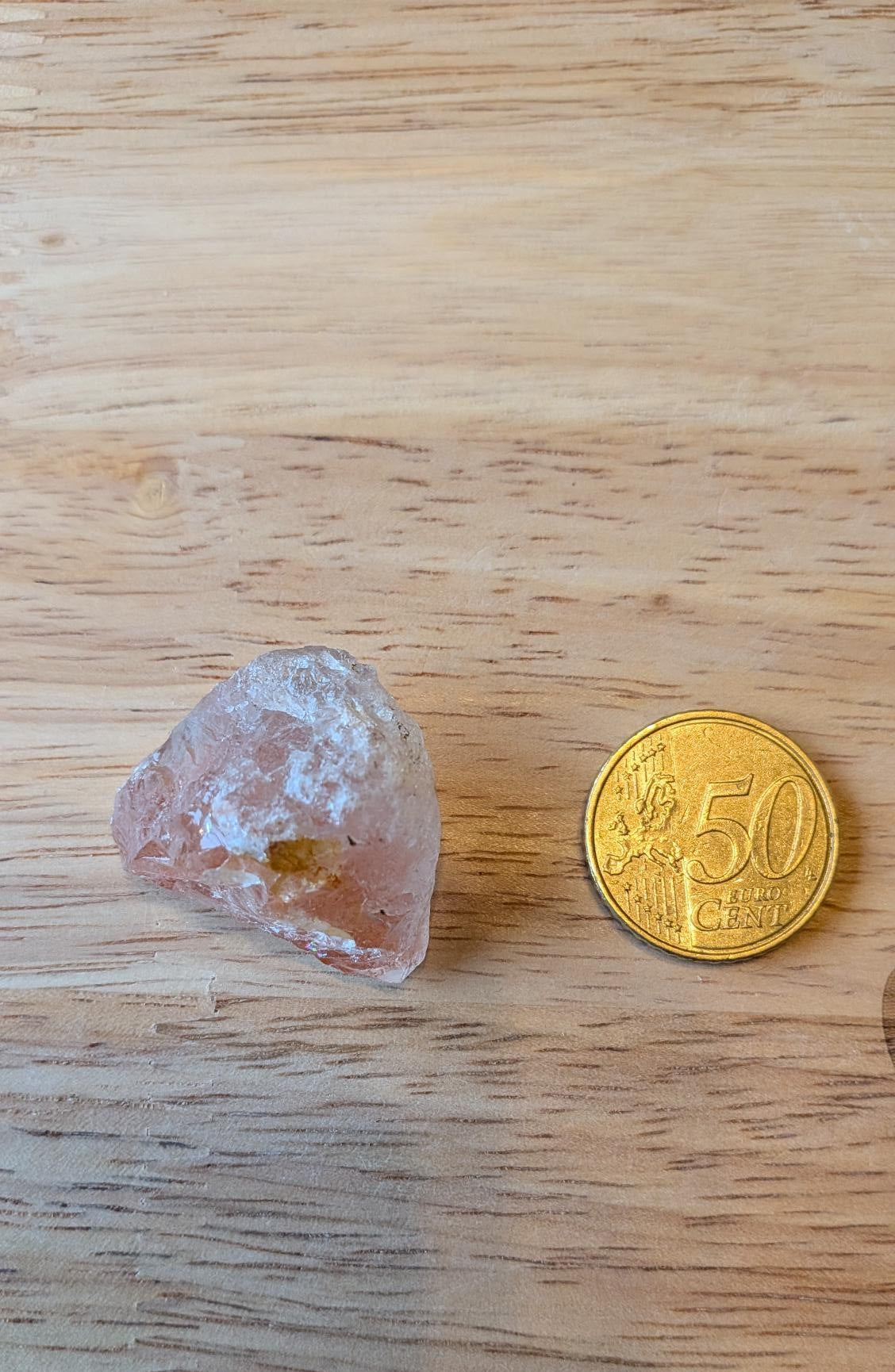 Een ruwe roze-oranje Helioliet edelsteen (Zonnesteen) op een lichte houten ondergrond naast een munt van 50 eurocent voor een custom wire wrapped hanger.