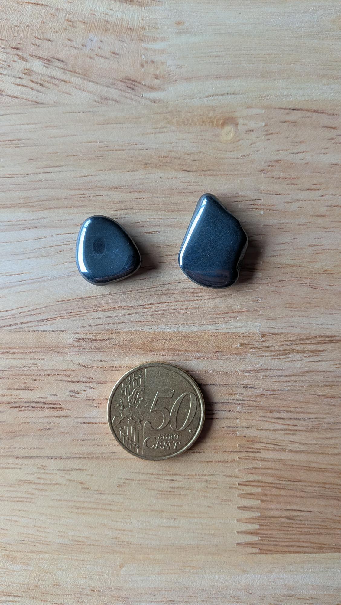 Twee glanzende, metallic grijze hematiet edelstenen in organische vormen naast een 50 eurocent munt op een houten ondergrond.