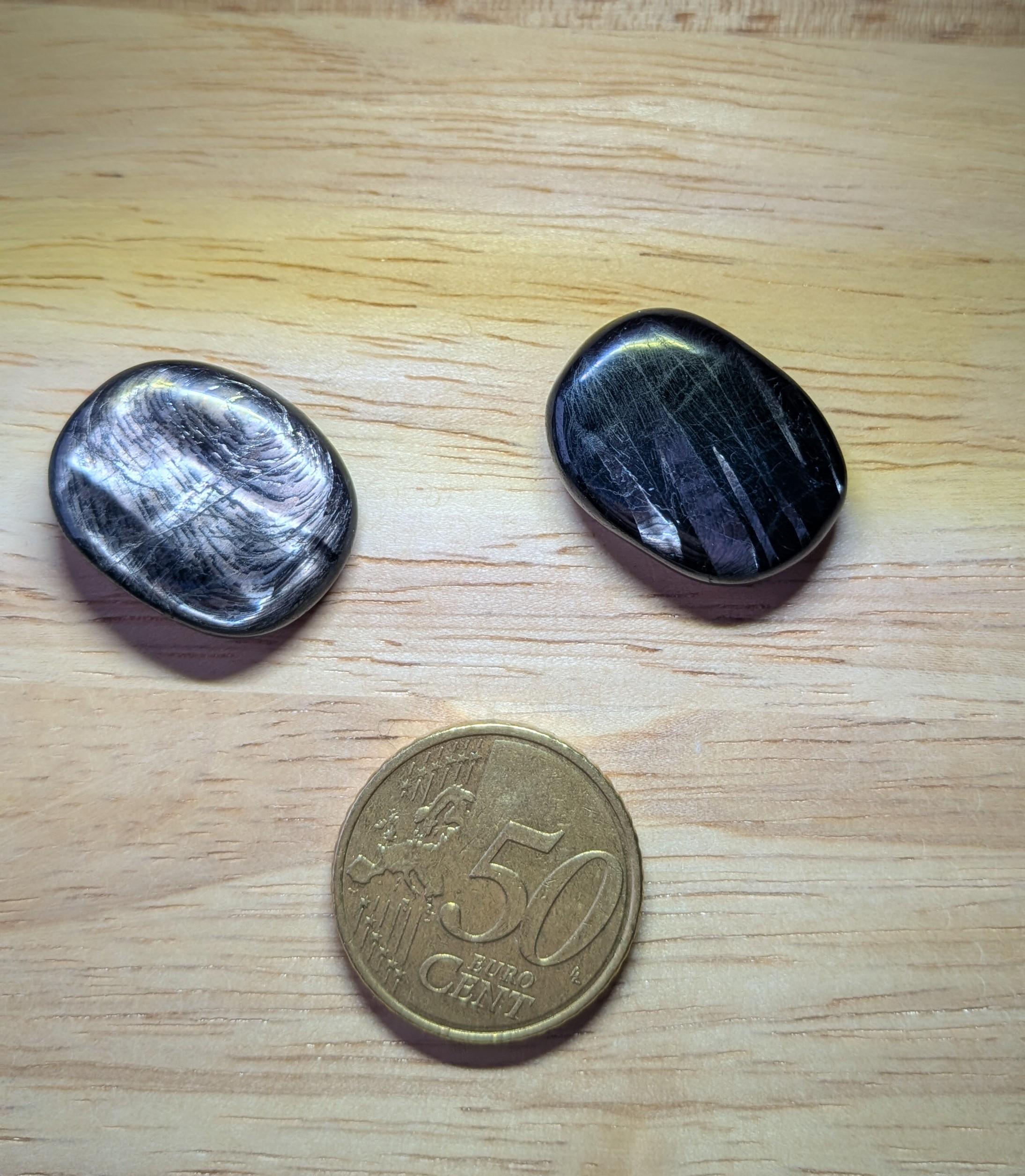 Twee gepolijste zwarte Hyperstheen edelstenen met een zilverachtige metaalglans naast een 50 eurocent munt.