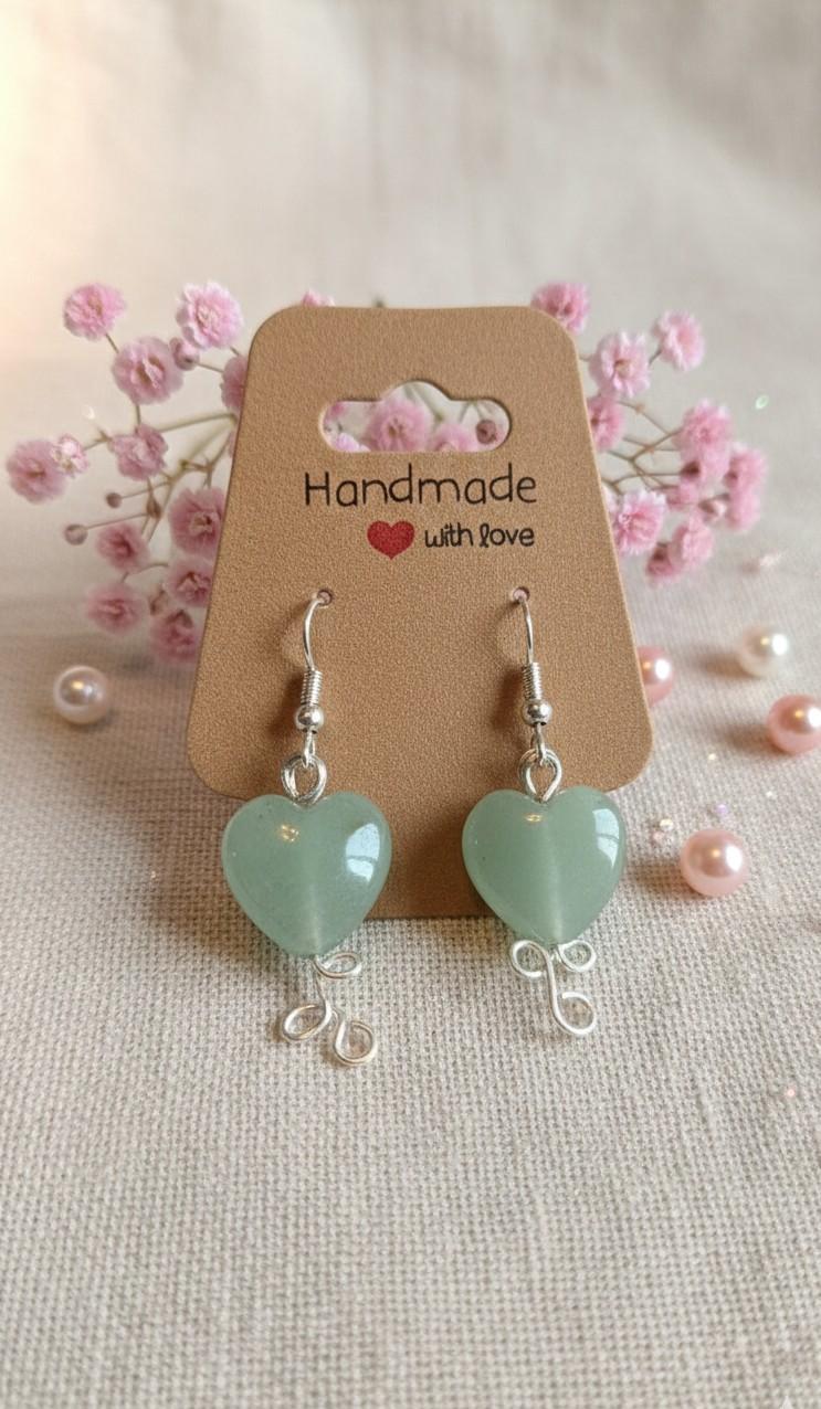 Handgemaakte zilverkleurige wire-wrap oorbellen met grote groene Jade hartjes, gepresenteerd op een "Handmade with love" kaartje met roze bloemetjes.