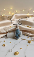 Handgemaakte verzilverde wire-wrap hanger met een blauw-glanzende Labradoriet edelsteen, gepresenteerd op een luxe witte marmeren ondergrond met goudkleurige accenten, champagnekleurige zijde en sfeervolle lichtjes.