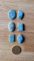 Zes natuurlijke Larimar edelstenen in verschillende vormen zoals ovaal en rechthoek op een houten ondergrond naast een 50 cent muntstuk.