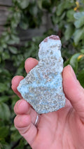 Prachtige gepolijste larimar plaat van 36 gram met unieke blauwe oceaan-tekening, vastgehouden in de hand tegen een achtergrond van groene bladeren.