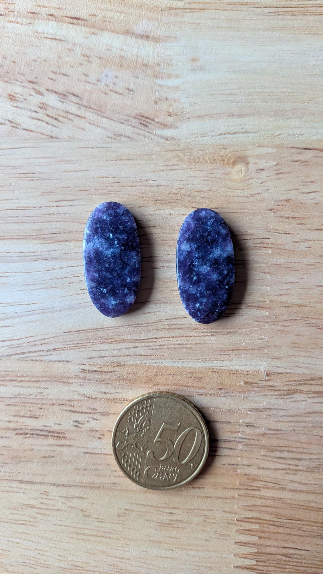 Twee ovale, gepolijste paarse lepidoliet edelstenen met natuurlijke glinsteringen naast een 50 eurocent munt op een houten achtergrond.