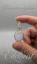 Handgemaakte wire wrapped hanger van Edeltwist met een ovale Regenboog Maansteen die een sterke blauwe flits toont, met de hand gewikkeld in sierlijk verzilverd draad tegen een neutrale, grijze studio-achtergrond.
