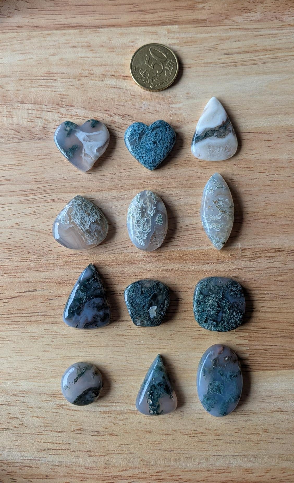 Twaalf verschillende vormen natuurlijke Mosagaat edelstenen met groene mosachtige insluitingen op een houten ondergrond naast een 50 cent muntstuk.
