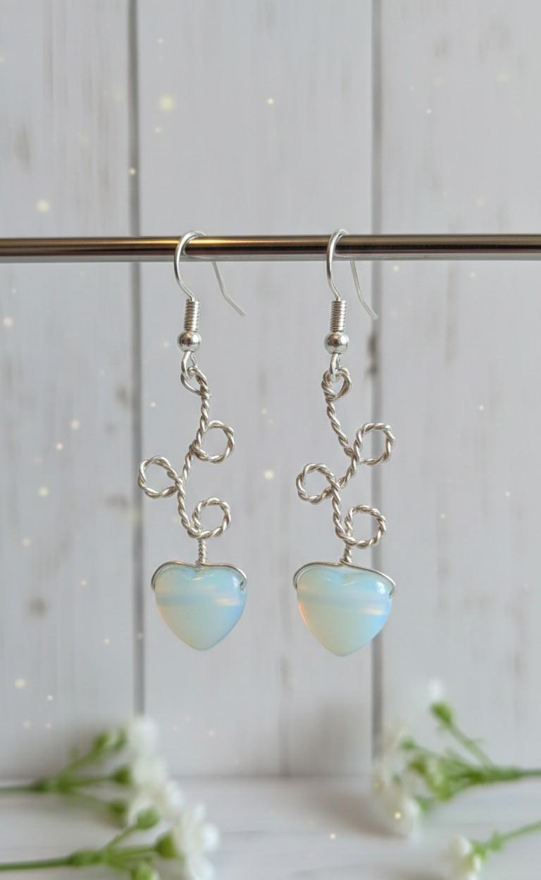 Handgemaakte zilverkleurige wire-wrap oorbellen met melkachtig witte Opaliet hartjes, gefotografeerd tegen een lichte achtergrond met witte bloemetjes.