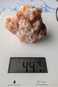 Een ruw, warm oranje Calciet blok van 447 gram, gefotografeerd in de hand en op een digitale weegschaal, met duidelijke natuurlijke kristalstructuren.