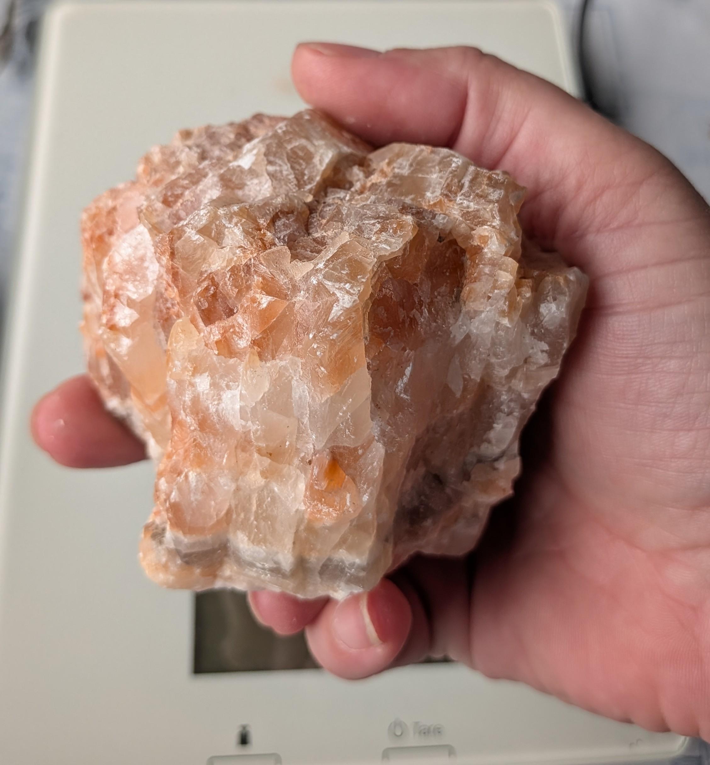 Een ruw, warm oranje Calciet blok van 447 gram, gefotografeerd in de hand en op een digitale weegschaal, met duidelijke natuurlijke kristalstructuren.