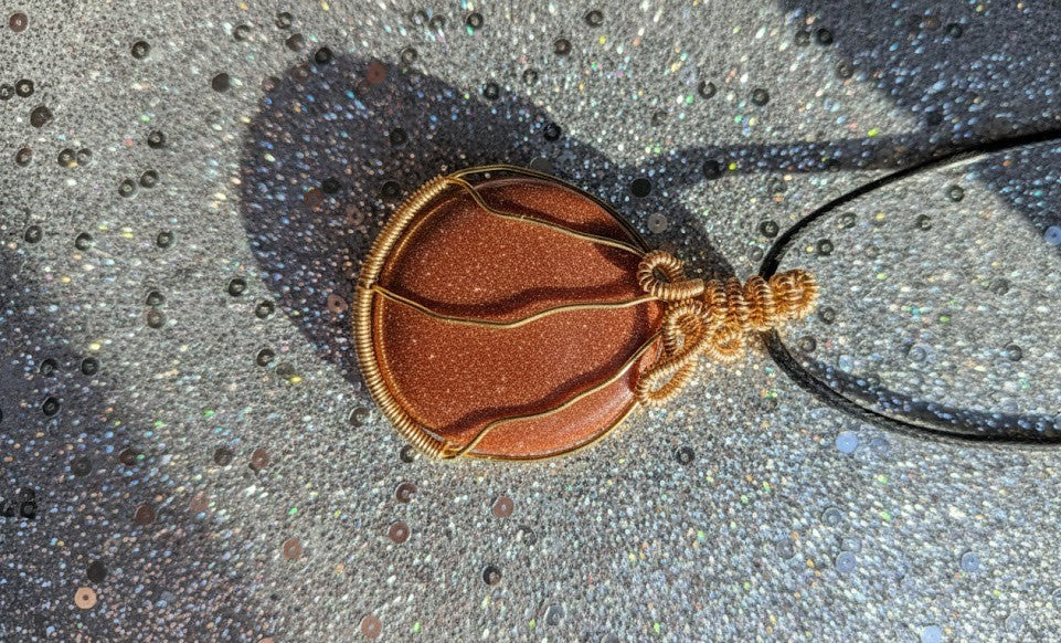 Close-up van een ronde, fonkelende oranje goldstone hanger, handgemaakt met koperkleurige wire wrapping techniek op een glitter achtergrond.