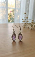 Gedetailleerde handgemaakte zilverkleurige oorbellen met een paarse fluoriet edelsteen, gewikkeld in fijn metaaldraad (wire-wrap), gefotografeerd tegen een lichte houten achtergrond.