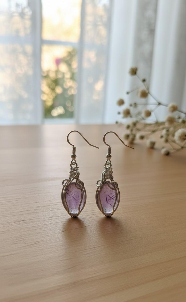 Gedetailleerde handgemaakte zilverkleurige oorbellen met een paarse fluoriet edelsteen, gewikkeld in fijn metaaldraad (wire-wrap), gefotografeerd tegen een lichte houten achtergrond.