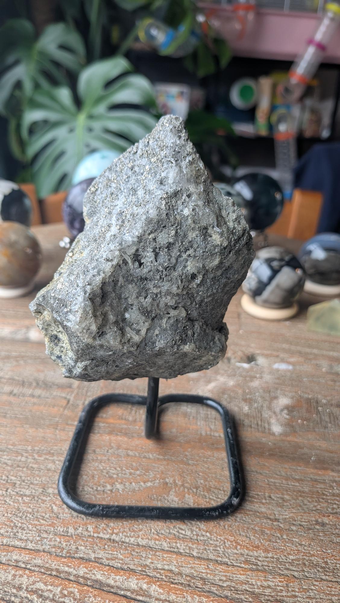 Ruwe grijze steen met goudkleurige pyriet glinsteringen en kwarts op een zwarte metalen standaard, item 215, 527 gram.