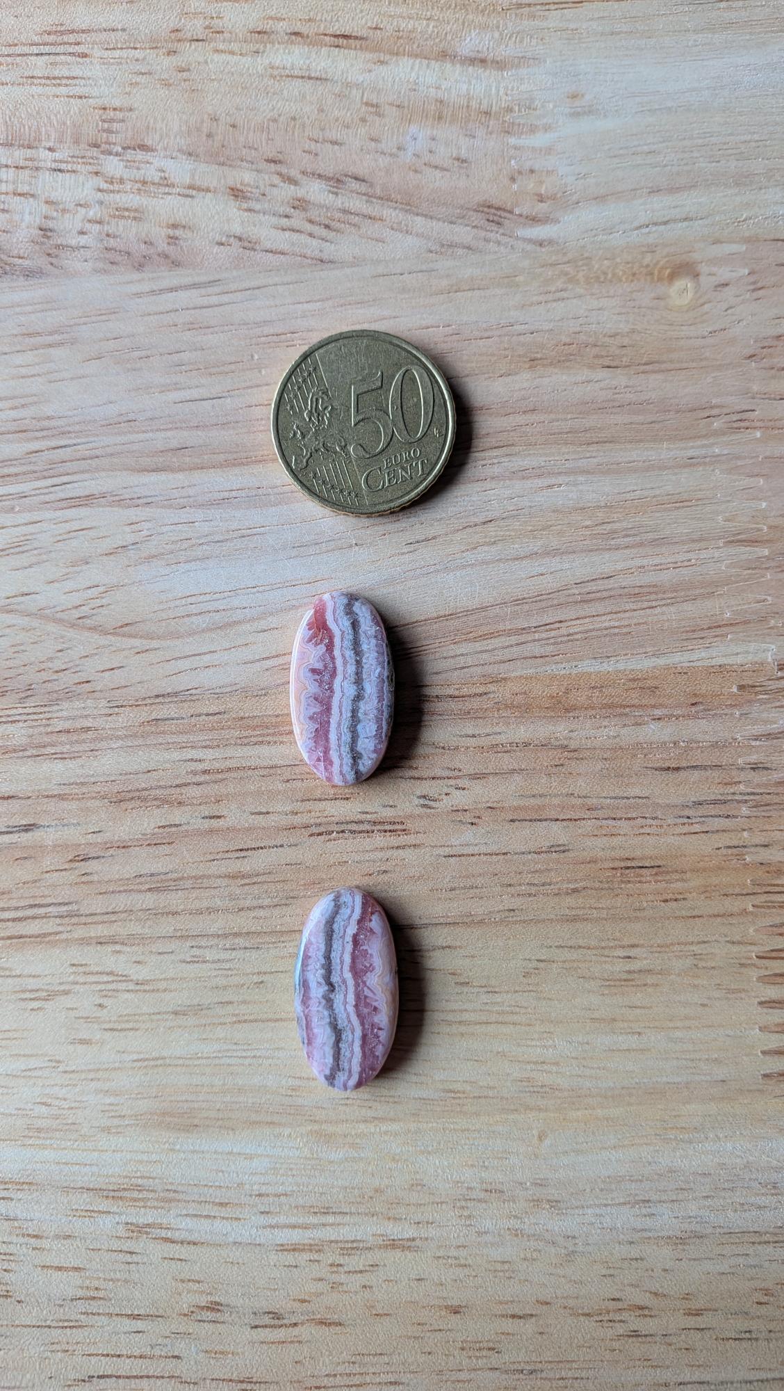 Twee ovale, gepolijste rhodochrosiet edelstenen met roze en witte bandenpatronen naast een 50 eurocent munt op een houten achtergrond.