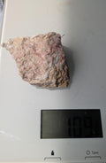Een ruw Rhyoliet-stuk van 109 gram met zachte roze-beige tinten en een korrelige textuur, gefotografeerd op een digitale weegschaal.