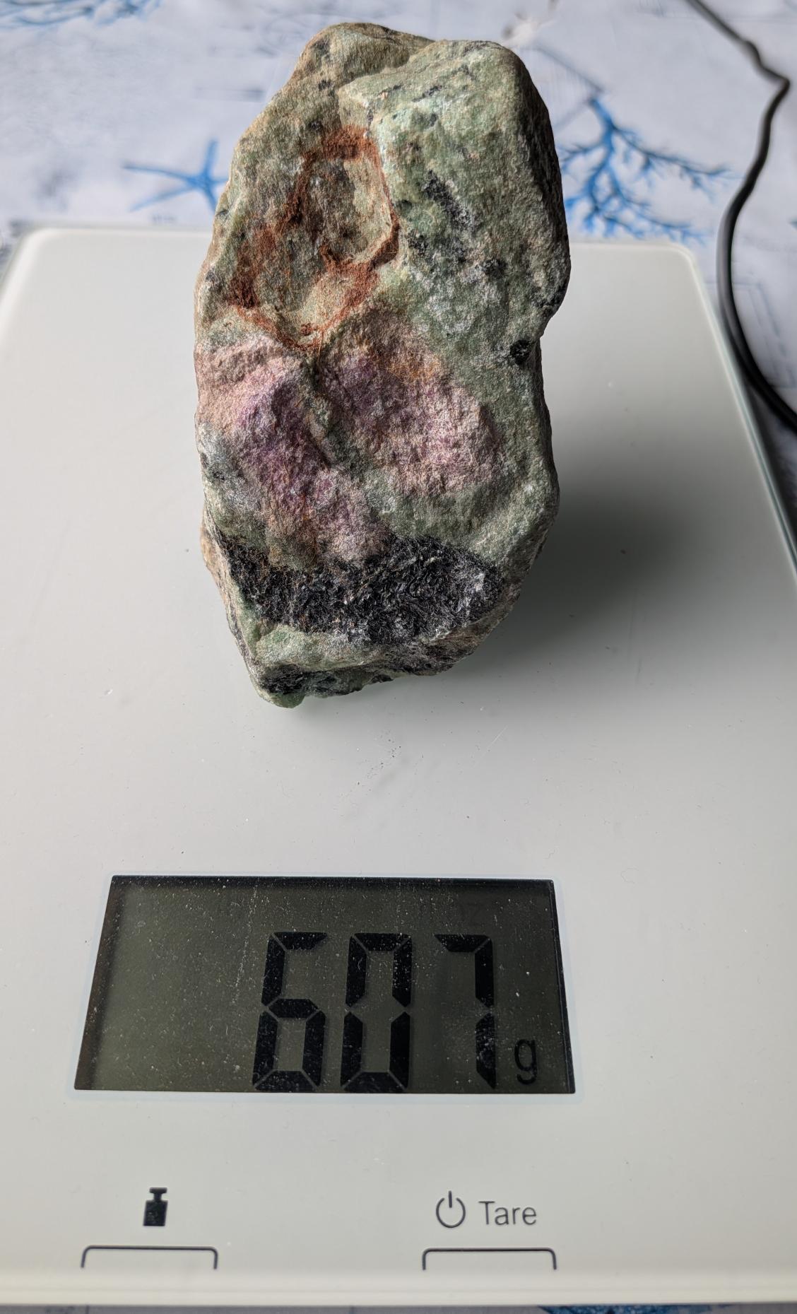 Een robuust, onbewerkt blok Robijn in Zoisiet van 607 gram, met opvallende roze robijn-insluitingen in een groen-zwart gestructureerd zoisiet-gesteente, gefotografeerd op een digitale weegschaal.