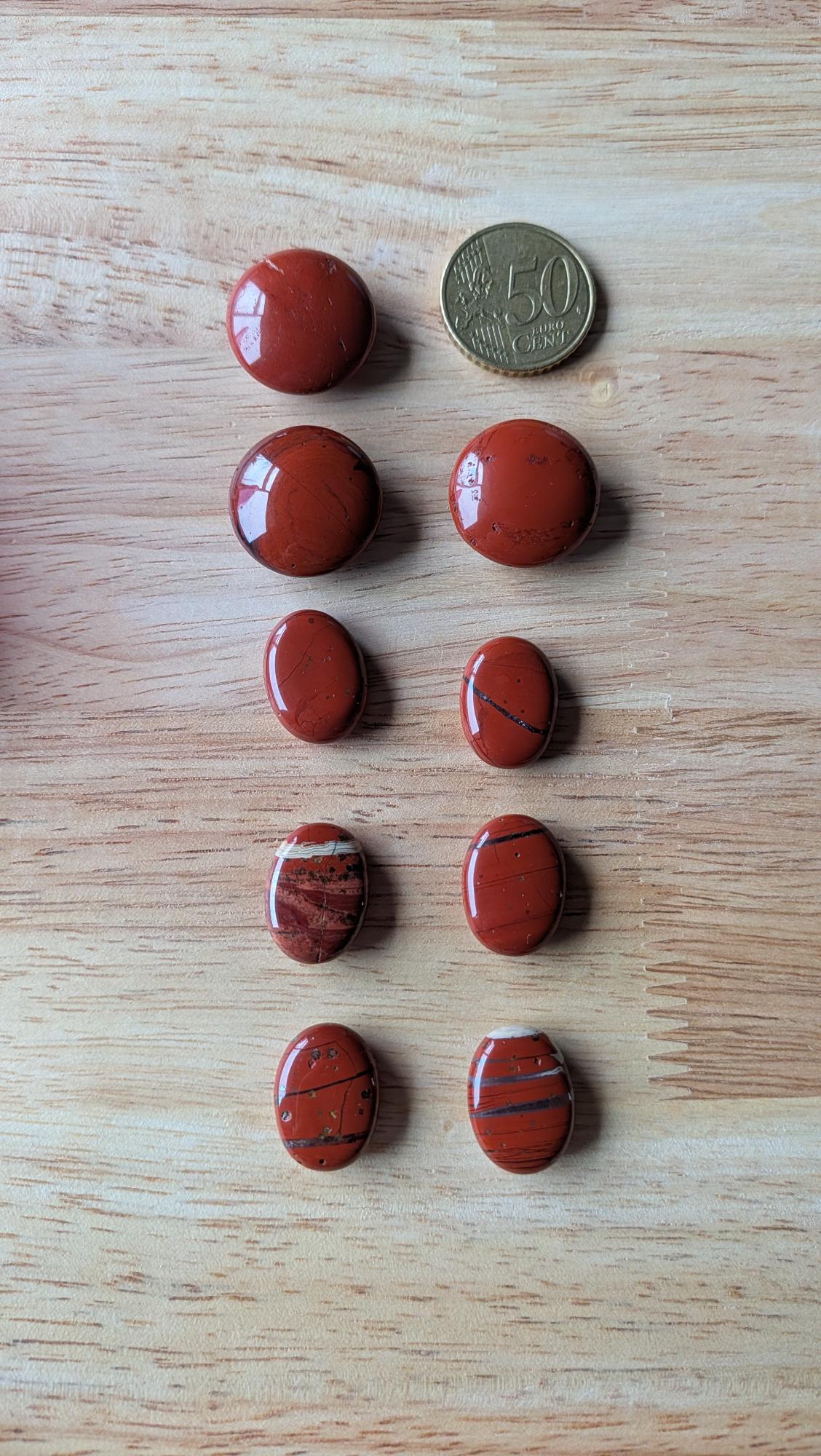 Negen gepolijste rode jaspis edelstenen in ronde en ovale vormen naast een 50 eurocent munt op een houten ondergrond voor een custom wire wrapped ontwerp.