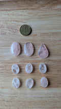 Negen gepolijste rozenkwarts edelstenen in ovale en druppelvormen naast een 50 eurocent munt op een houten achtergrond.