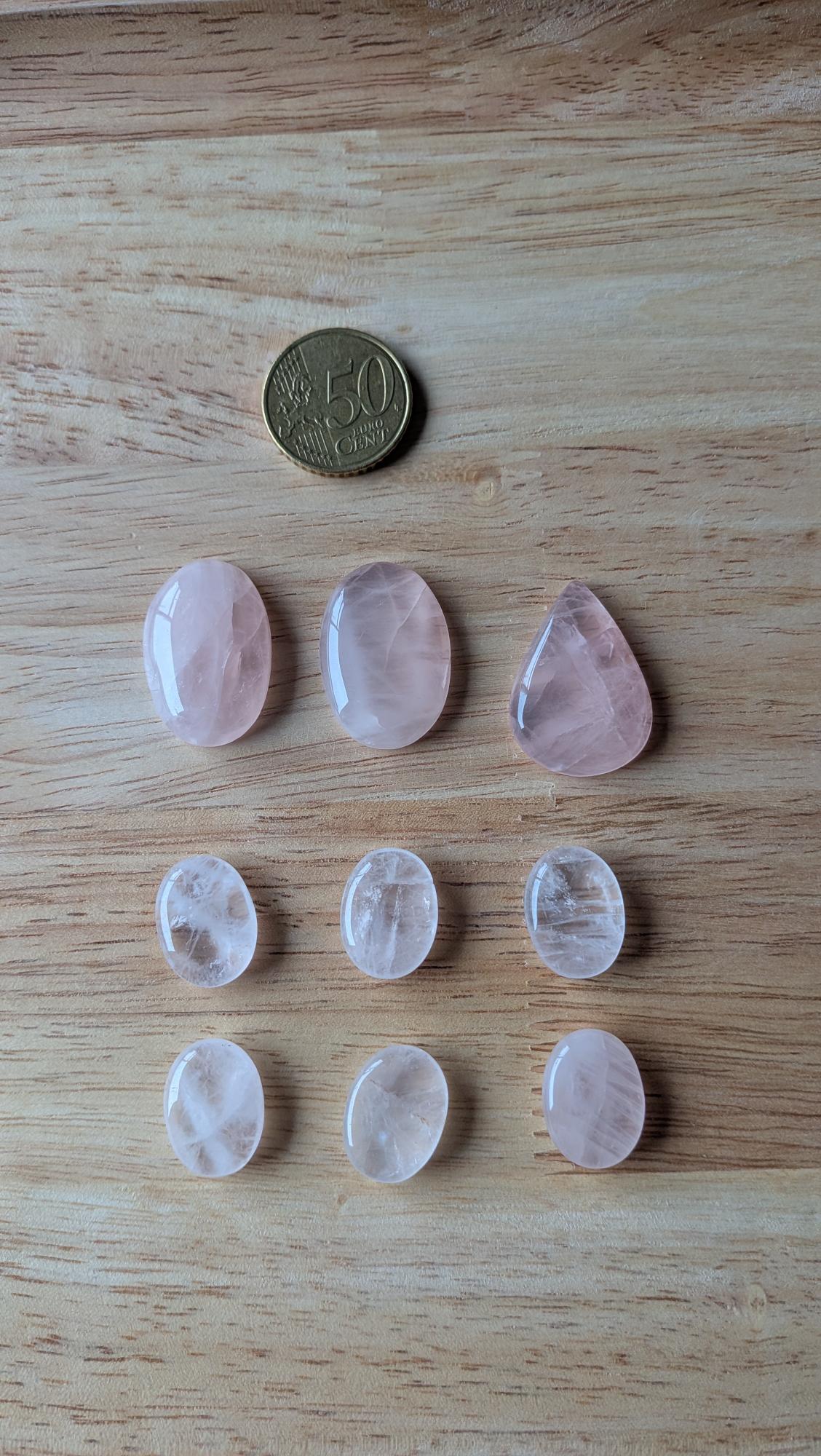 Negen gepolijste rozenkwarts edelstenen in ovale en druppelvormen naast een 50 eurocent munt op een houten achtergrond.
