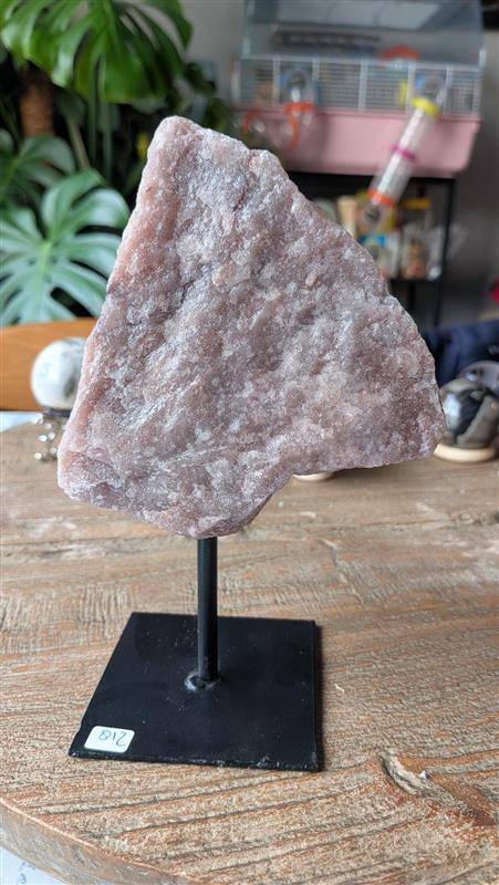 Breed vooraanzicht van een grote, ruwe roze rozenkwarts edelsteen op een zwarte metalen standaard. De steen weegt 702 gram en toont een natuurlijke, kristalachtige textuur.