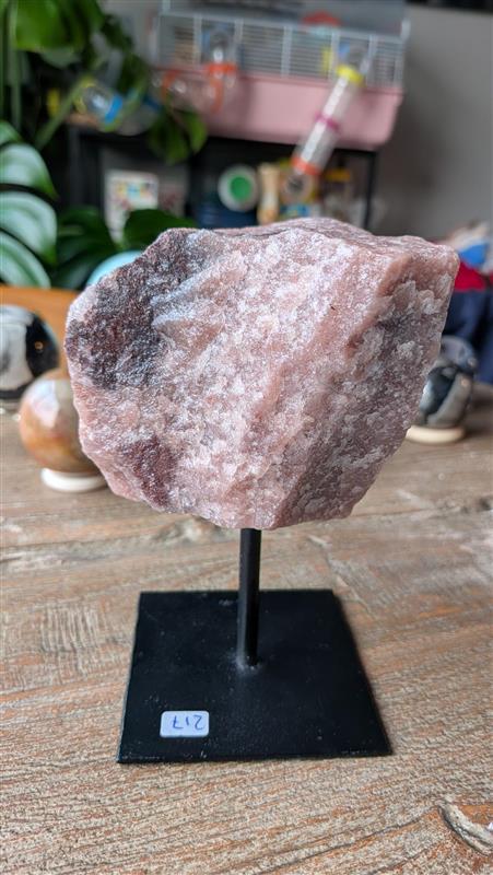 Zijaanzicht van een ruwe, roze rozenkwarts edelsteen gemonteerd op een zwarte metalen standaard. De steen heeft een grove textuur en een natuurlijke, donkere verkleuring aan de linkerzijde.