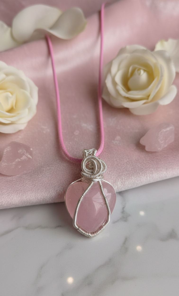 Handgemaakte zilverkleurige wire-wrapped hanger met een hartvormige Rozenkwarts aan een roze koord, gepresenteerd op een luxe roze zijden achtergrond met witte rozen.