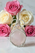 Handgemaakte verzilverde wire-wrapped hanger met een ronde roze rozenkwarts edelsteen, gepresenteerd tegen een achtergrond van zachte roze en witte rozen op een marmeren ondergrond.